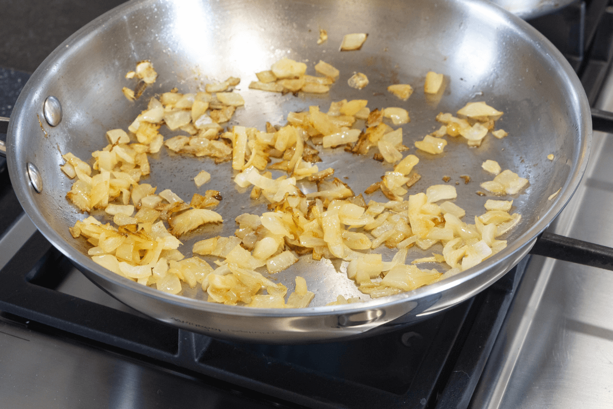 Cebolas refogadas e douradas soltas no fundo de uma frigideira de inox brilhante, exemplificando a estética e a facilidade de limpeza da cozinha antiaderente.