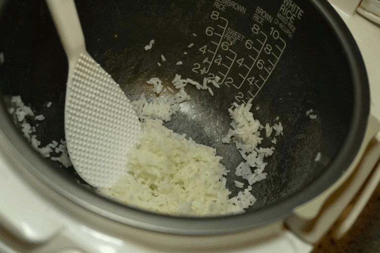 Interior da cuba de uma panela elétrica com arroz branco cozido e soltinho, sendo mexido com a espátula de plástico própria para não riscar.