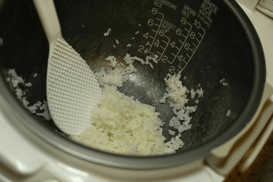 Interior da cuba de uma panela elétrica com arroz branco cozido e soltinho, sendo mexido com a espátula de plástico própria para não riscar.