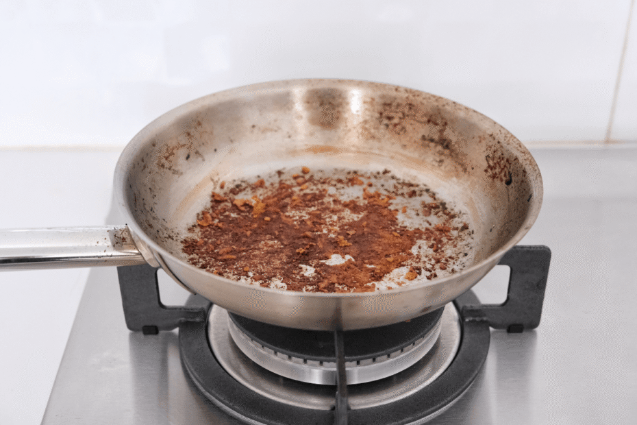 Frigideira de aço inox sobre o fogão com resíduos de alimentos queimados e grudados no fundo, ilustrando a necessidade de aprender como fazer uma frigideira ficar antiaderente corretamente.