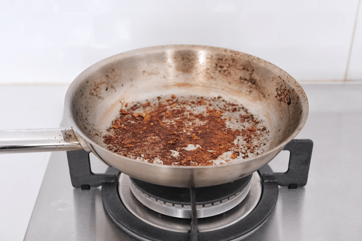 Frigideira de aço inox sobre o fogão com resíduos de alimentos queimados e grudados no fundo, ilustrando a necessidade de aprender como fazer uma frigideira ficar antiaderente corretamente.