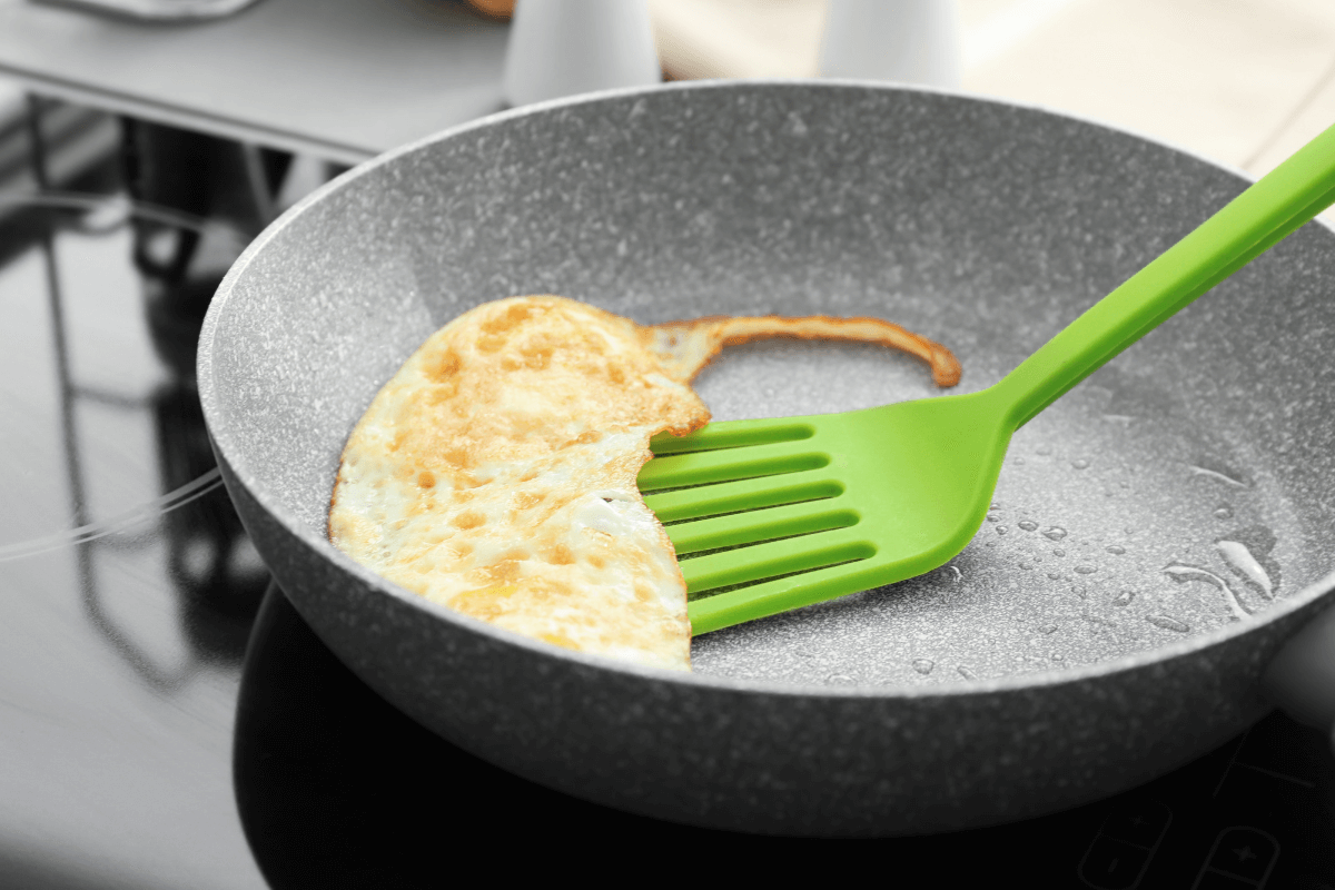 Espátula de silicone verde virando um ovo frito em frigideira com revestimento cinza, demonstrando o uso de utensílios macios que não riscam o fundo da panela.
