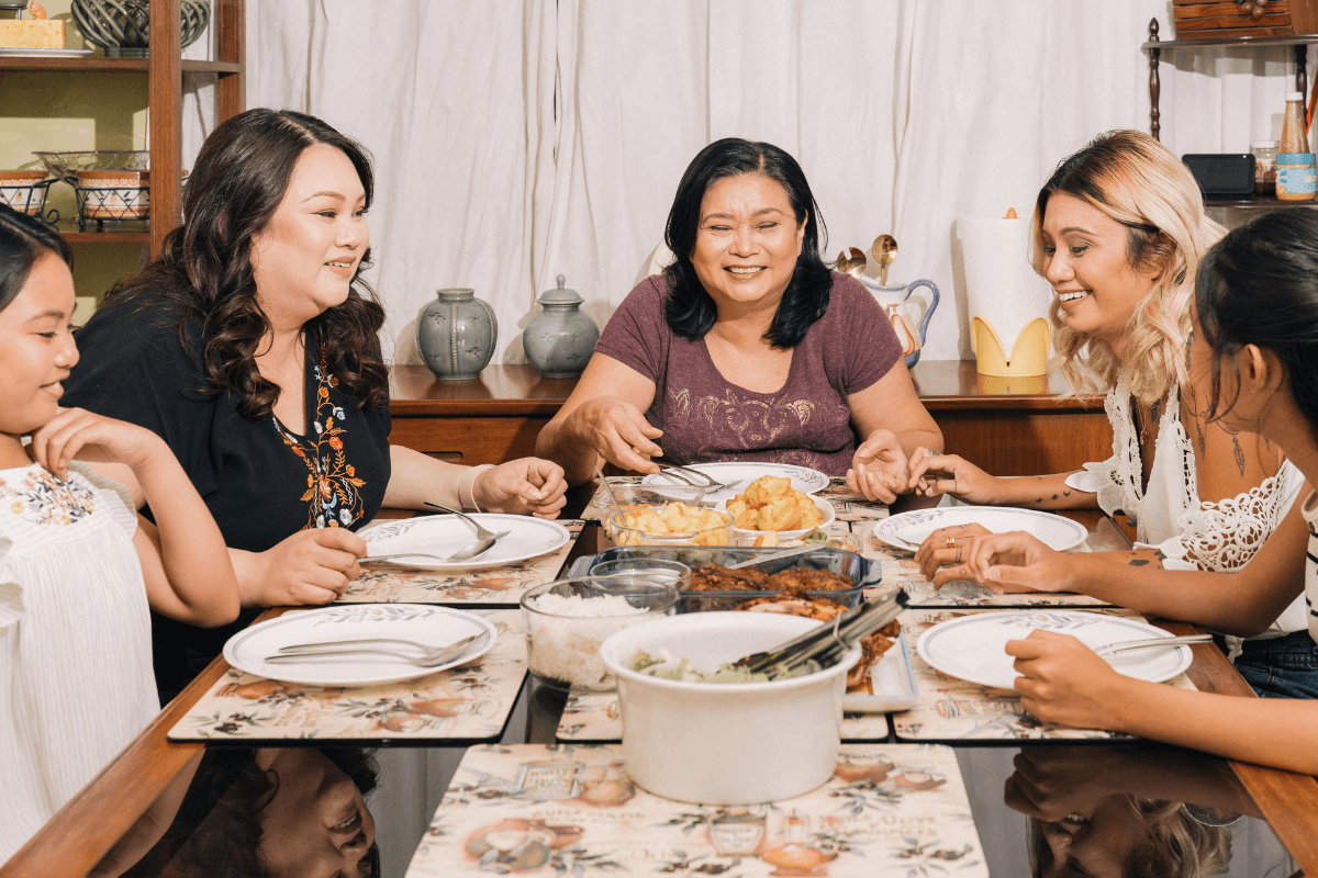 Família numerosa de três gerações reunida sorrindo à mesa de jantar servida com arroz e acompanhamentos, ilustrando o benefício da panela elétrica para rotinas onde várias pessoas comem em horários próximos.