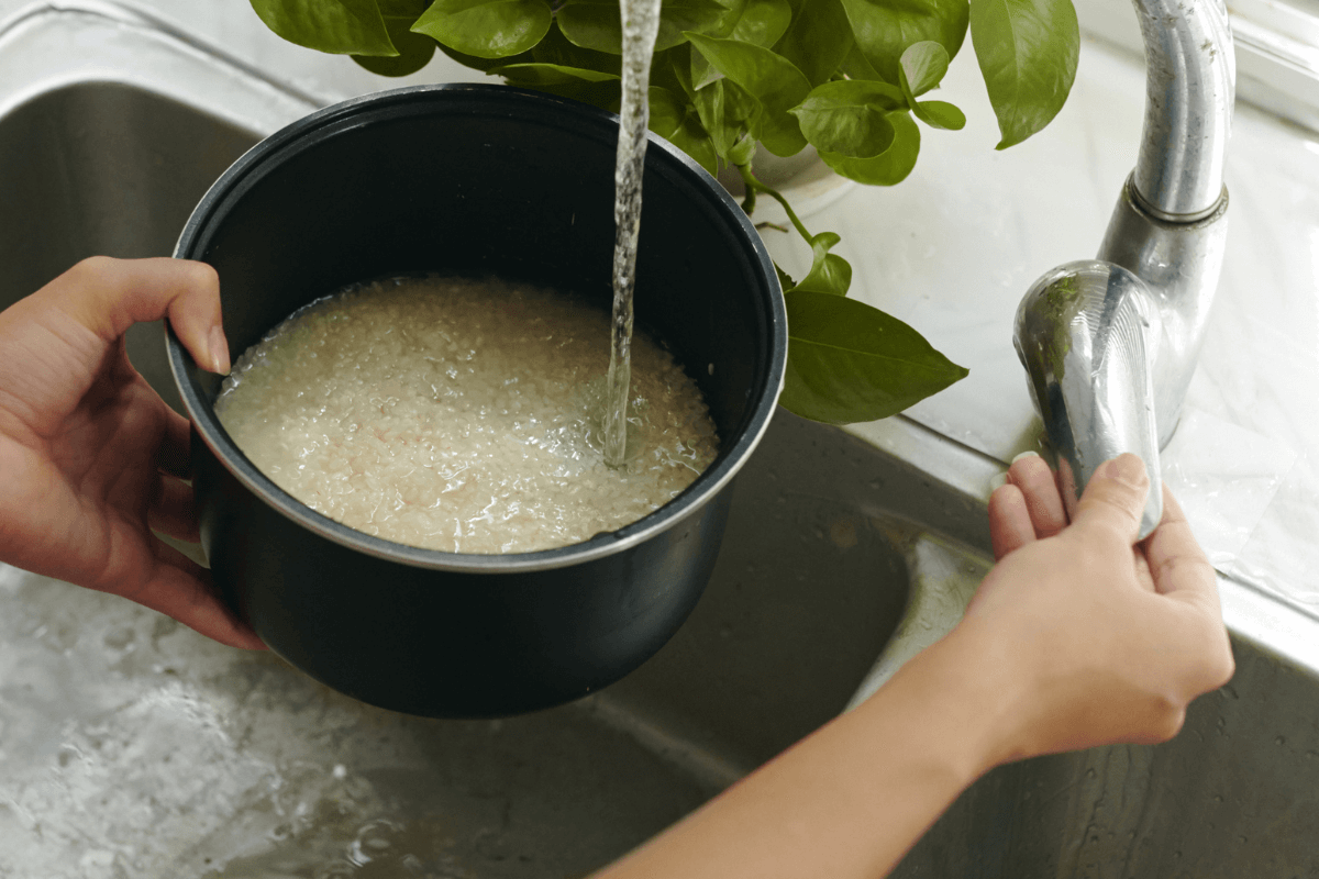 Mãos lavando arroz dentro da cuba antiaderente da panela elétrica sob a torneira da cozinha, demonstrando o preparo prático antes do cozimento.