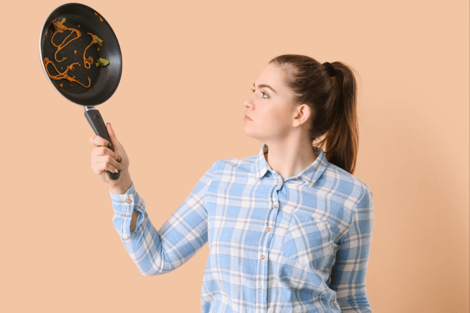 Mulher com expressão frustrada segurando uma frigideira antiaderente suja com restos de comida grudados, ilustrando o resultado de não saber a técnica correta de untar.