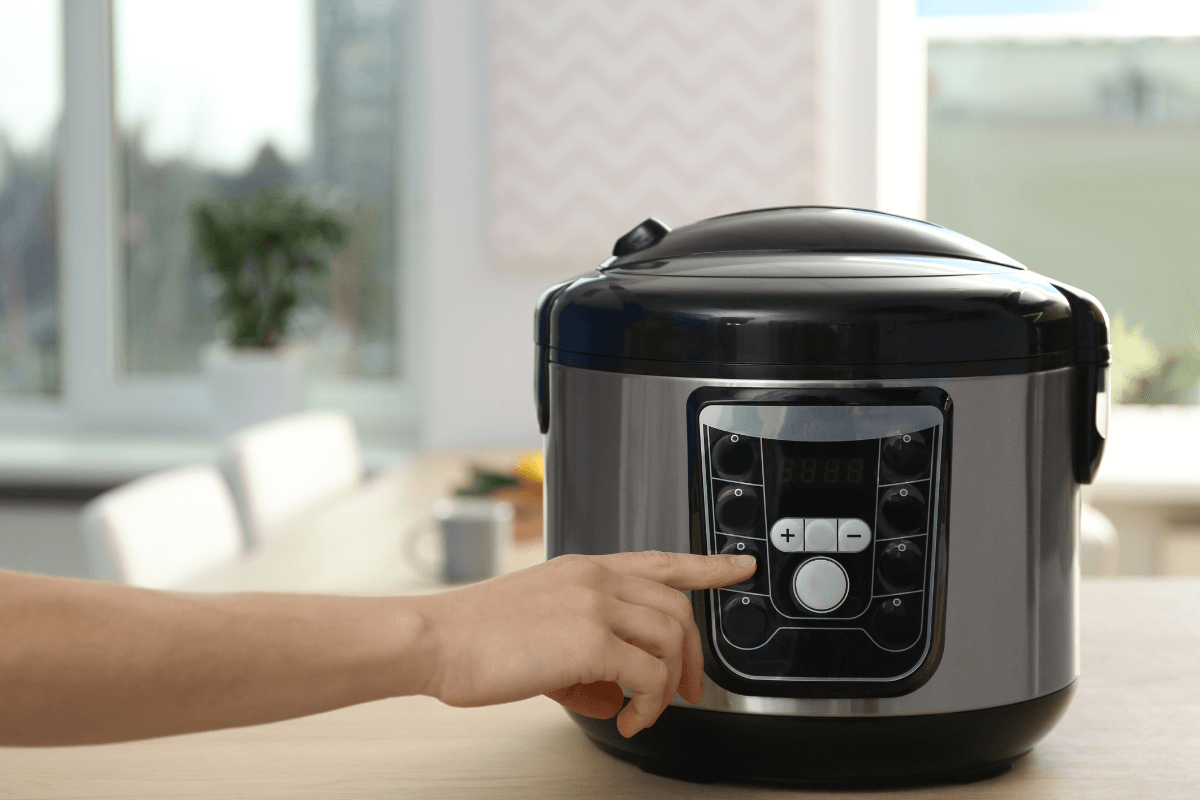Detalhe de uma mão ajustando o painel digital de uma panela de arroz elétrica em inox, demonstrando a facilidade de programação do equipamento moderno.