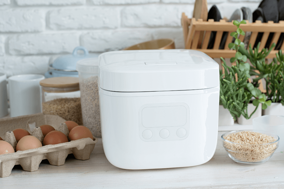 Panela de arroz elétrica branca com tampa fechada sobre a bancada, ilustrando a segurança de cozinhar sem fogo exposto ao lado de ingredientes frescos.