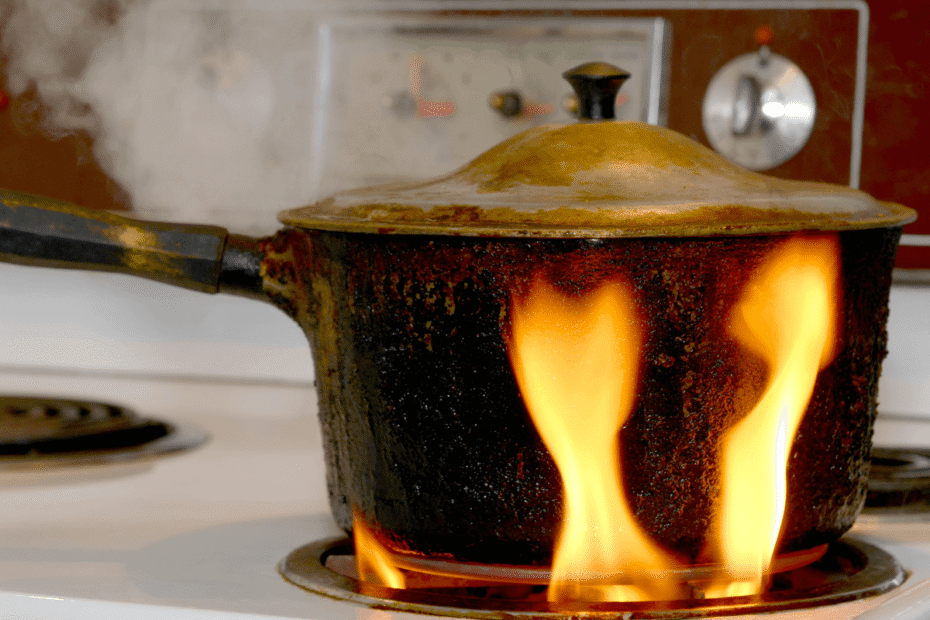 Panela muito velha e queimada no fogão com chamas altas e fumaça, mostrando o perigo de usar utensílios desgastados que soltam resíduos na comida.