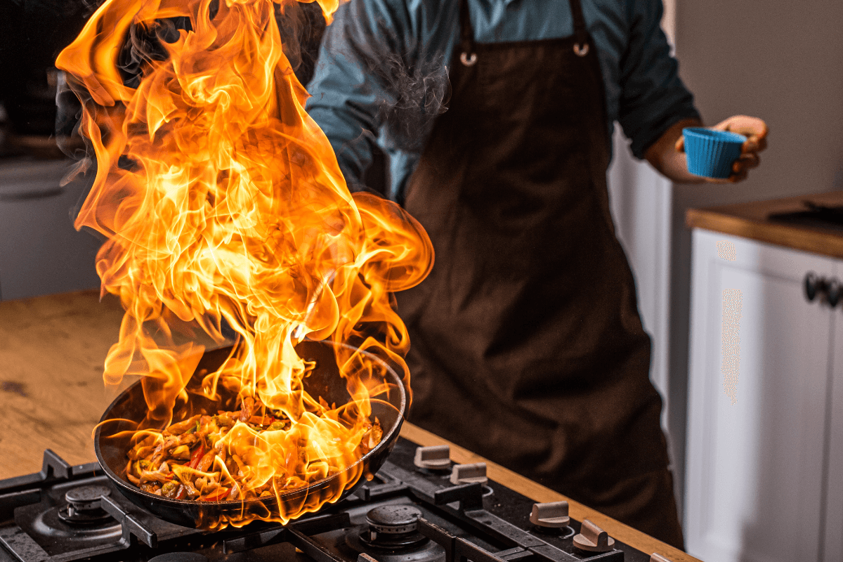 Chef segurando uma frigideira em chamas no fogão, ilustrando o superaquecimento extremo que antecede o choque térmico e causa a deformação do metal.